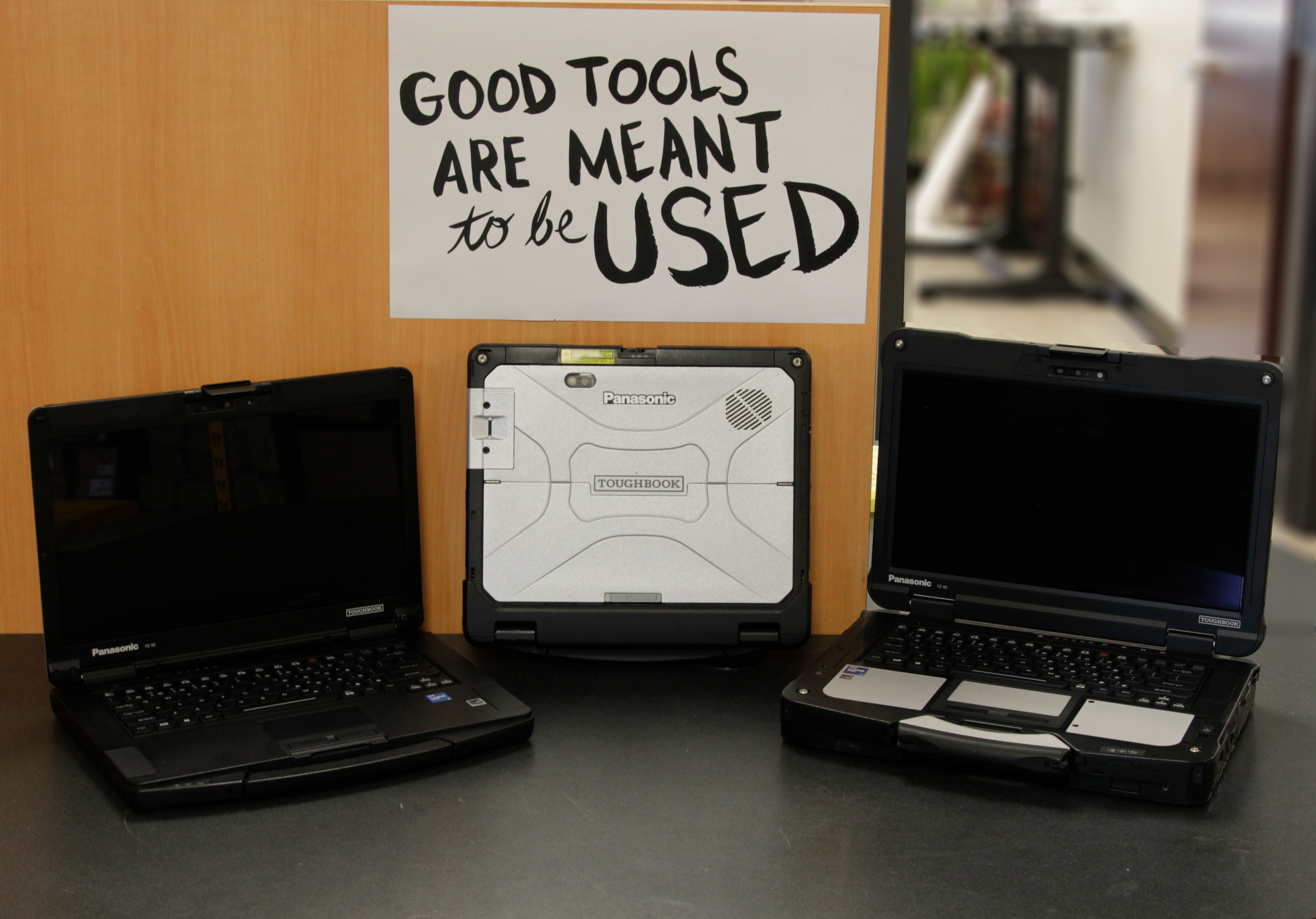 A Panasonic Toughbook FZ-40, FZ-55 and CF-33 sitting in front of a Good Tools Are Meant to Be Used Poster